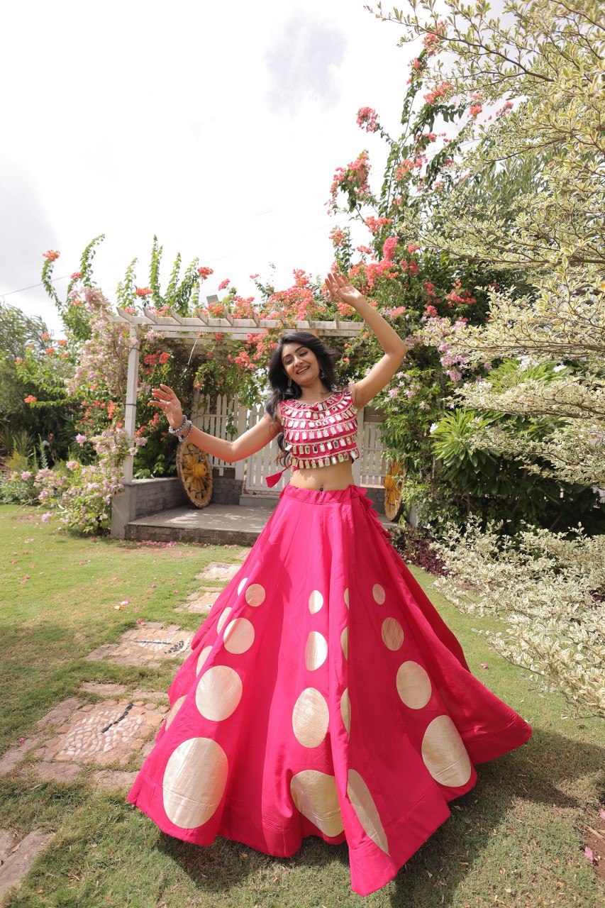 Woman in a pink and white polka dot outfit standing in a garden with flowers and a pergola.