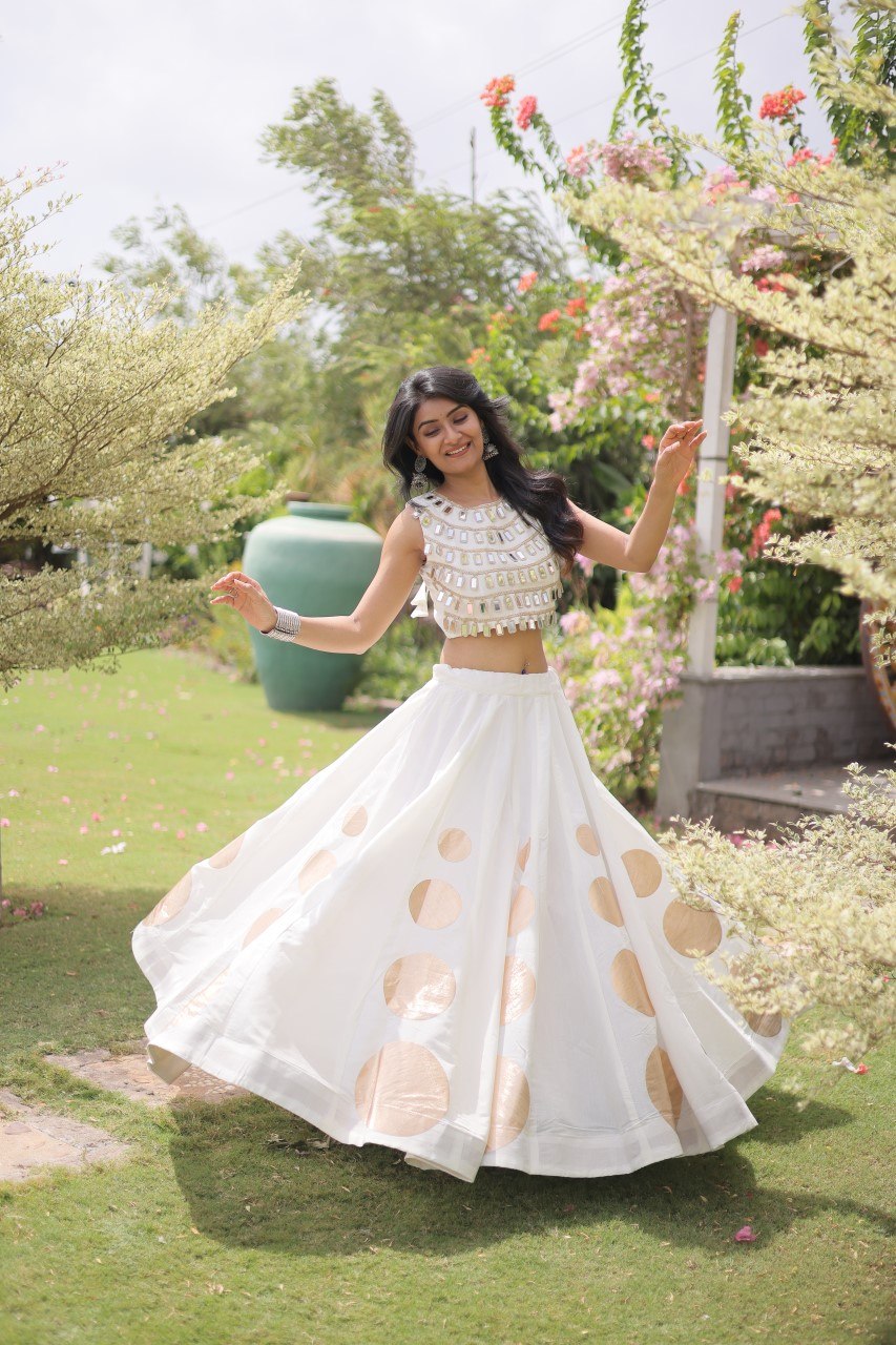 Woman in a white and gold traditional outfit standing in a garden with flowers and greenery.