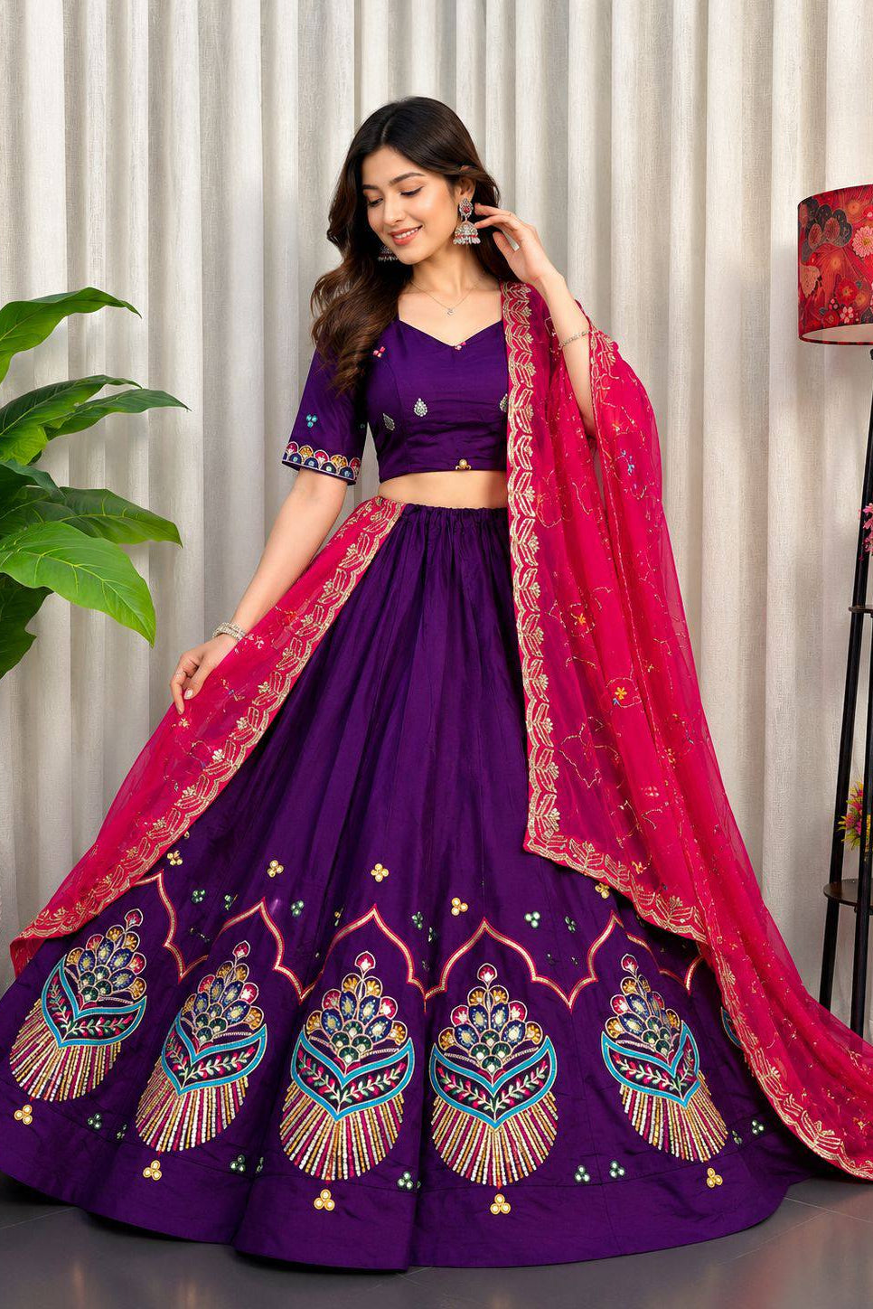 A woman models a traditional Indian outfit consisting of a purple lehenga skirt with colorful embroidered motifs, a matching purple crop top, and a bright pink dupatta with gold borders.
