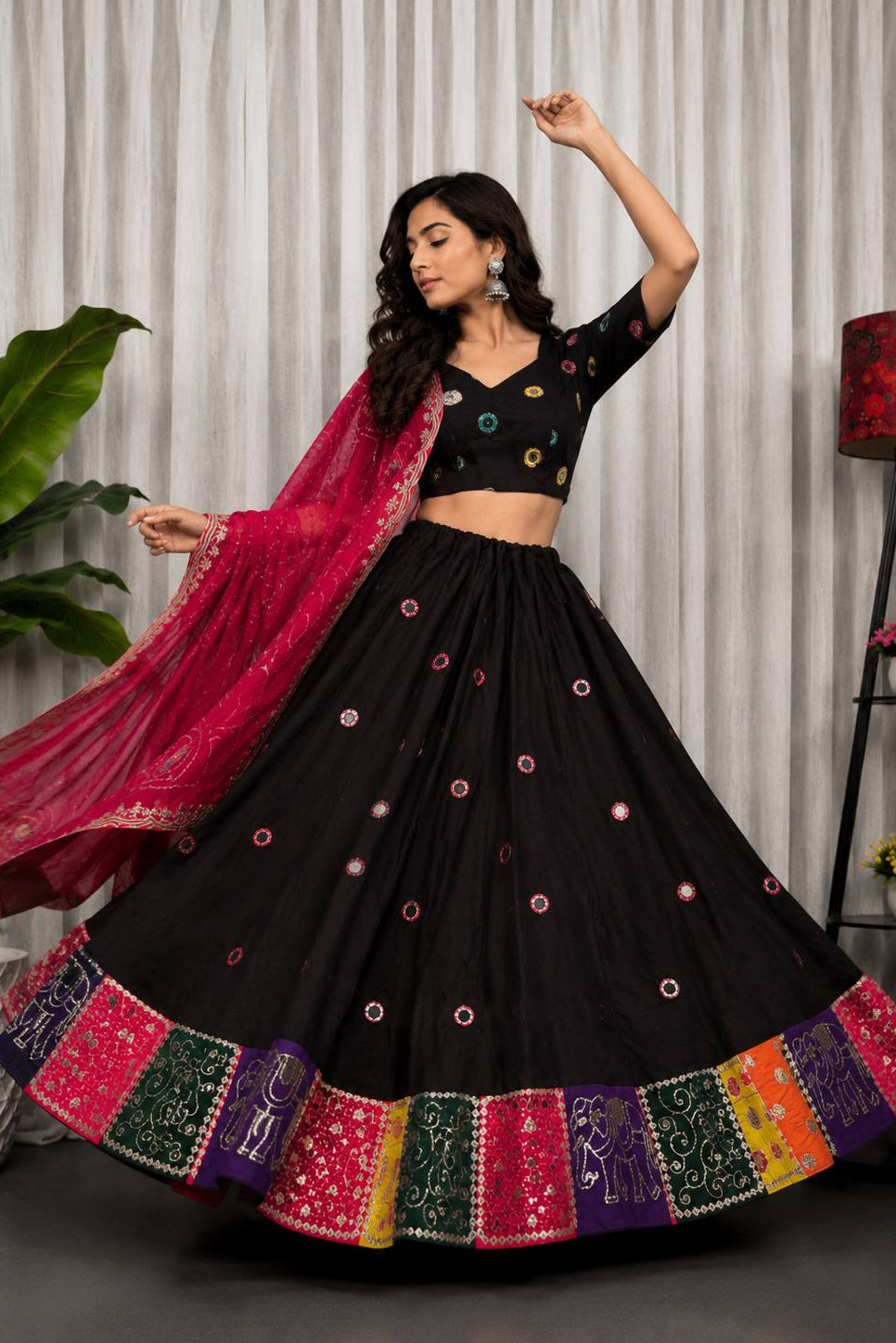 A woman modeling a black embroidered lehenga choli set with a bright pink dupatta, posed in front of a neutral curtain backdrop with decorative plants and a lamp.