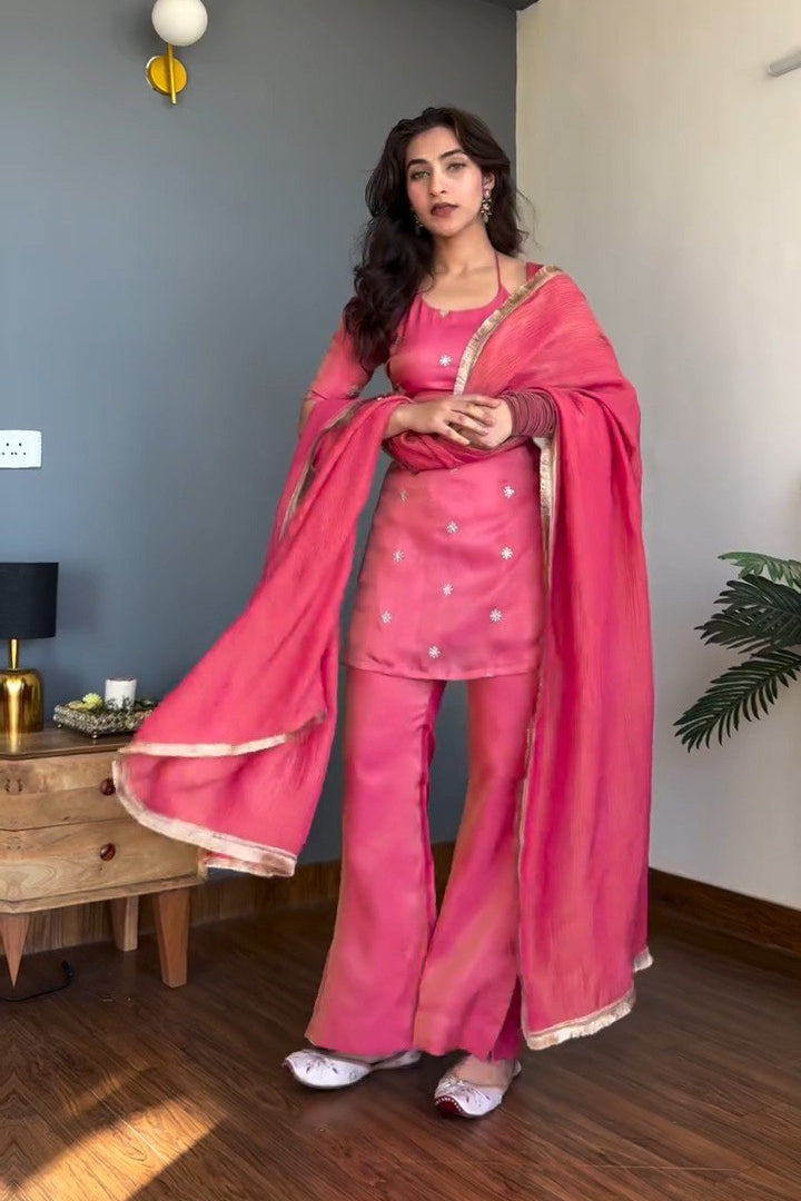 Woman in a pink traditional outfit standing in a room with a gray wall and wooden floor.