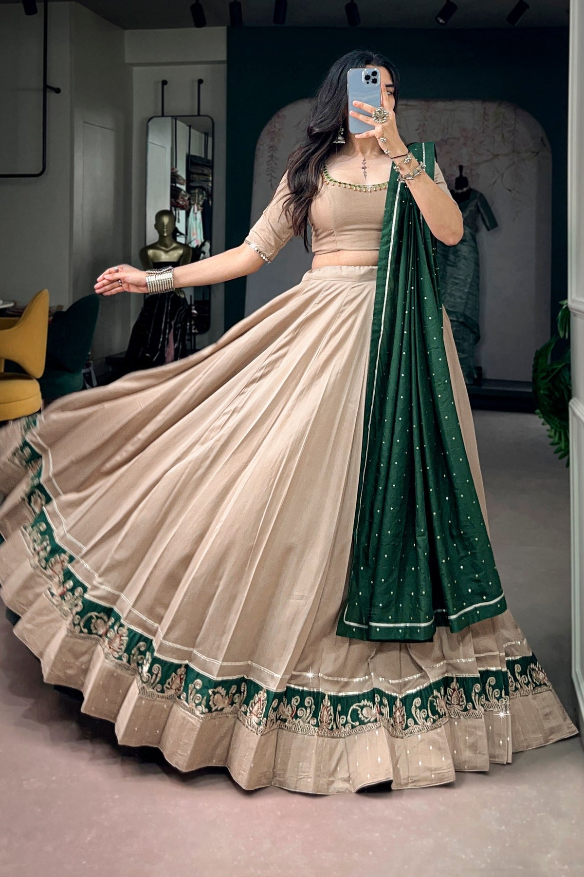 A woman wearing a beige and green traditional Indian lehenga choli set with a matching dupatta, taking a mirror selfie in an indoor boutique setting.