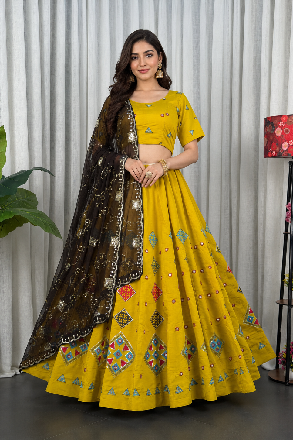 A woman models a bright yellow Indian ethnic lehenga choli set with a dark brown embroidered dupatta, standing in front of a white curtain backdrop.