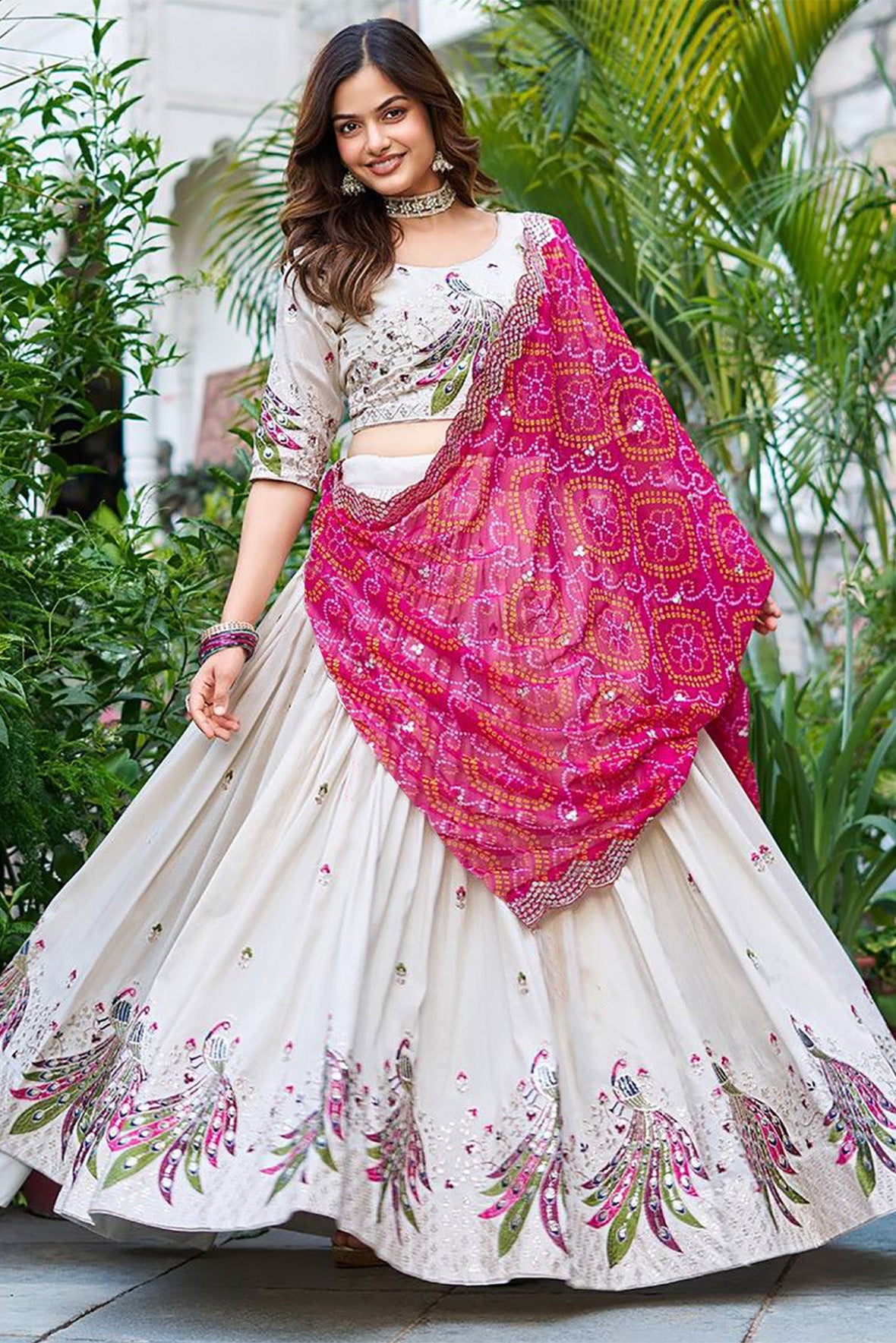Woman in a traditional outfit with a pink and white patterned dupatta, standing outdoors with greenery.