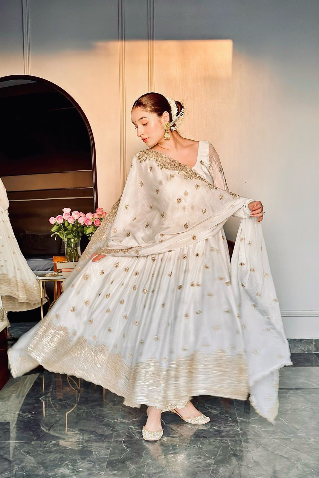 Woman in a white traditional outfit with gold details standing in a room with a mirror and flowers.