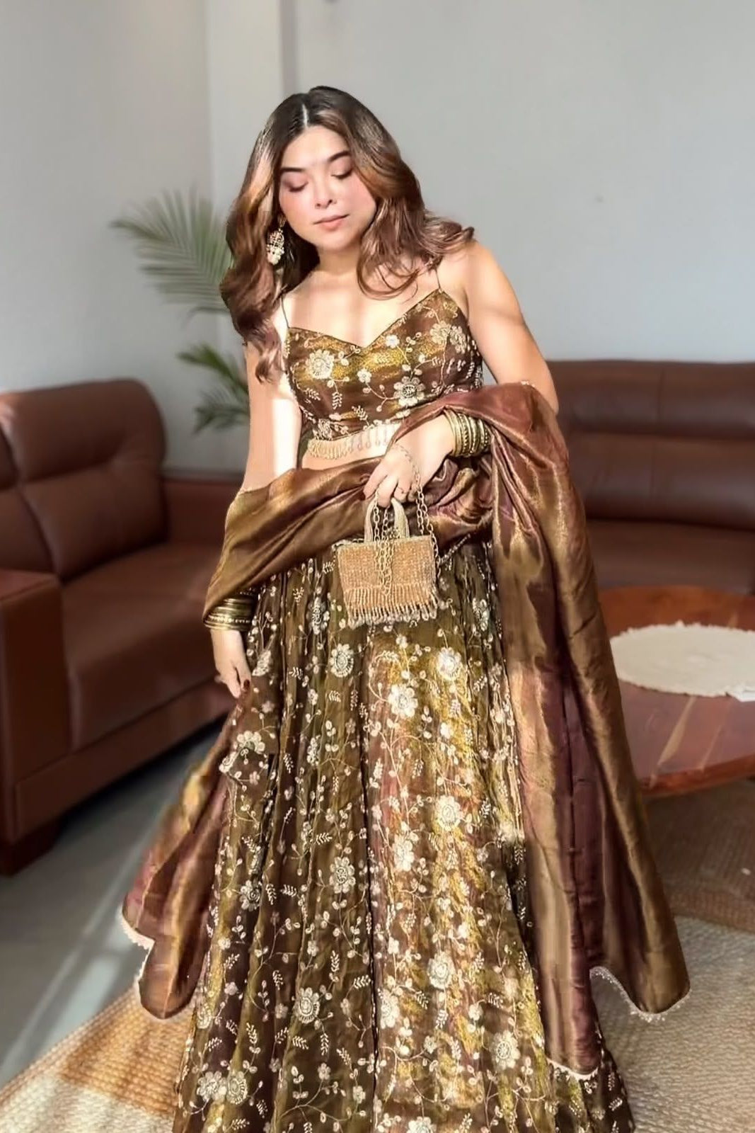 A woman models a brown and gold floral lehenga choli set with a matching dupatta in a living room setting.