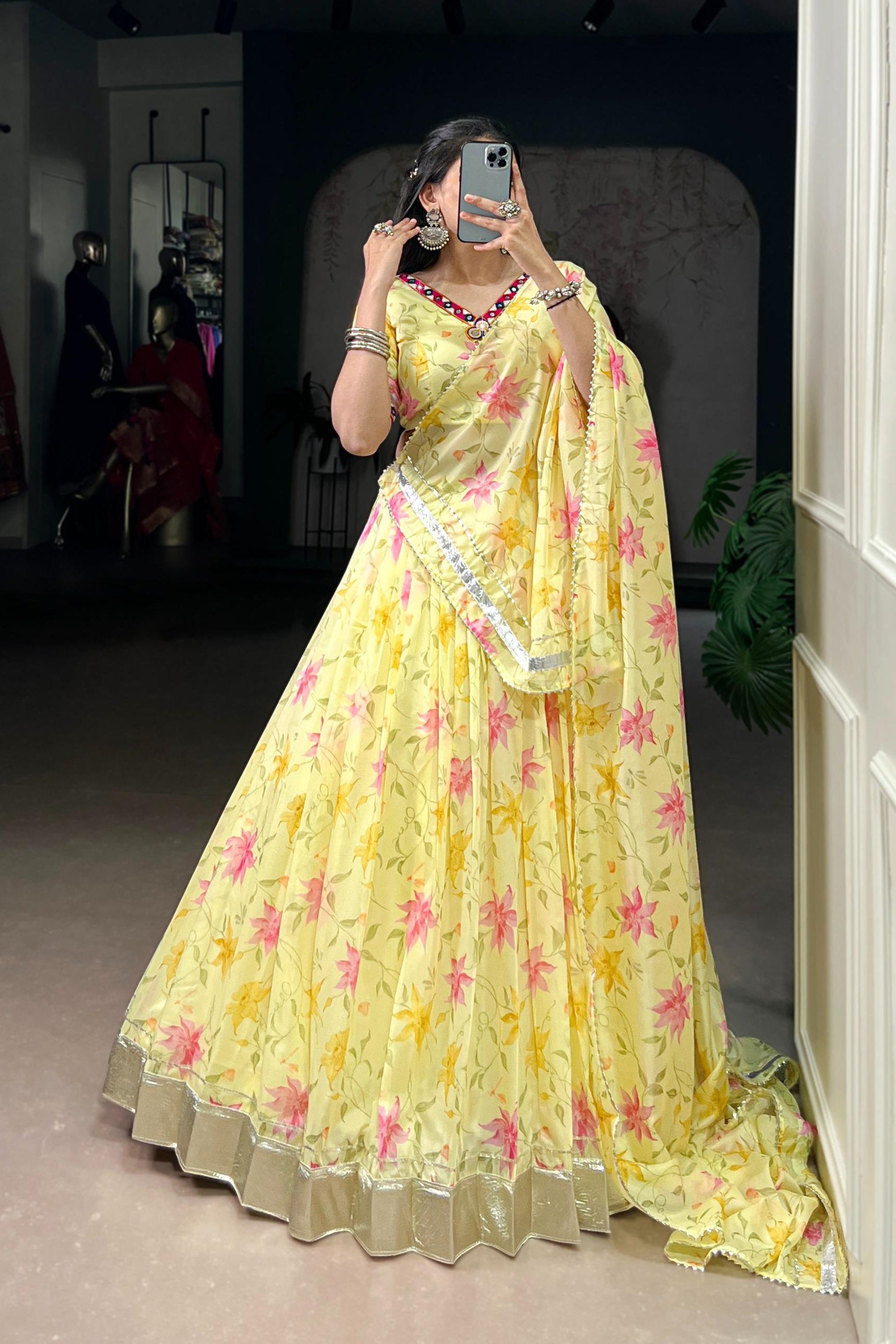 A woman takes a mirror selfie wearing a yellow floral lehenga choli set with pink and yellow flowers, in what appears to be a boutique showroom.