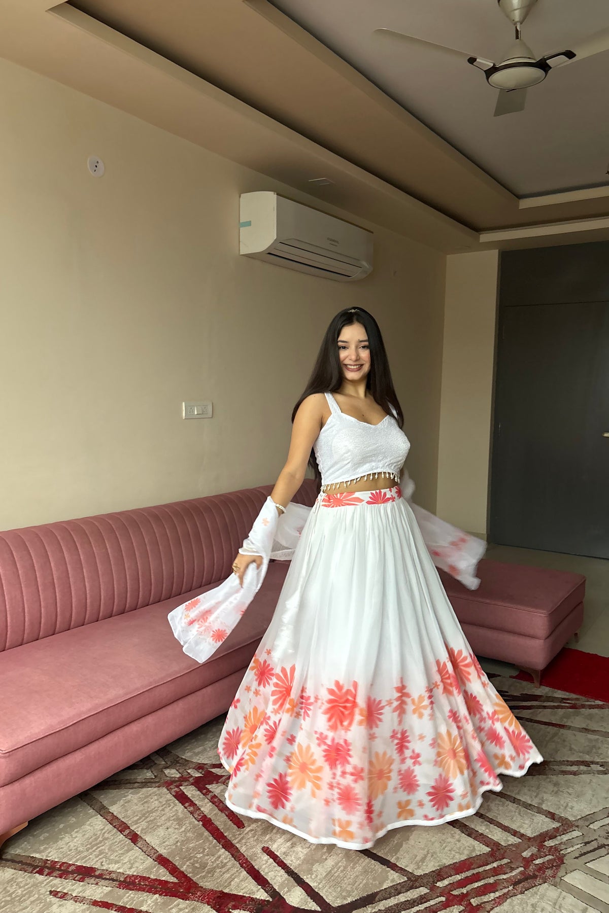 A woman modeling a white sleeveless lehenga choli set with a coral and orange floral print on the skirt, standing in a living room with a pink sofa.