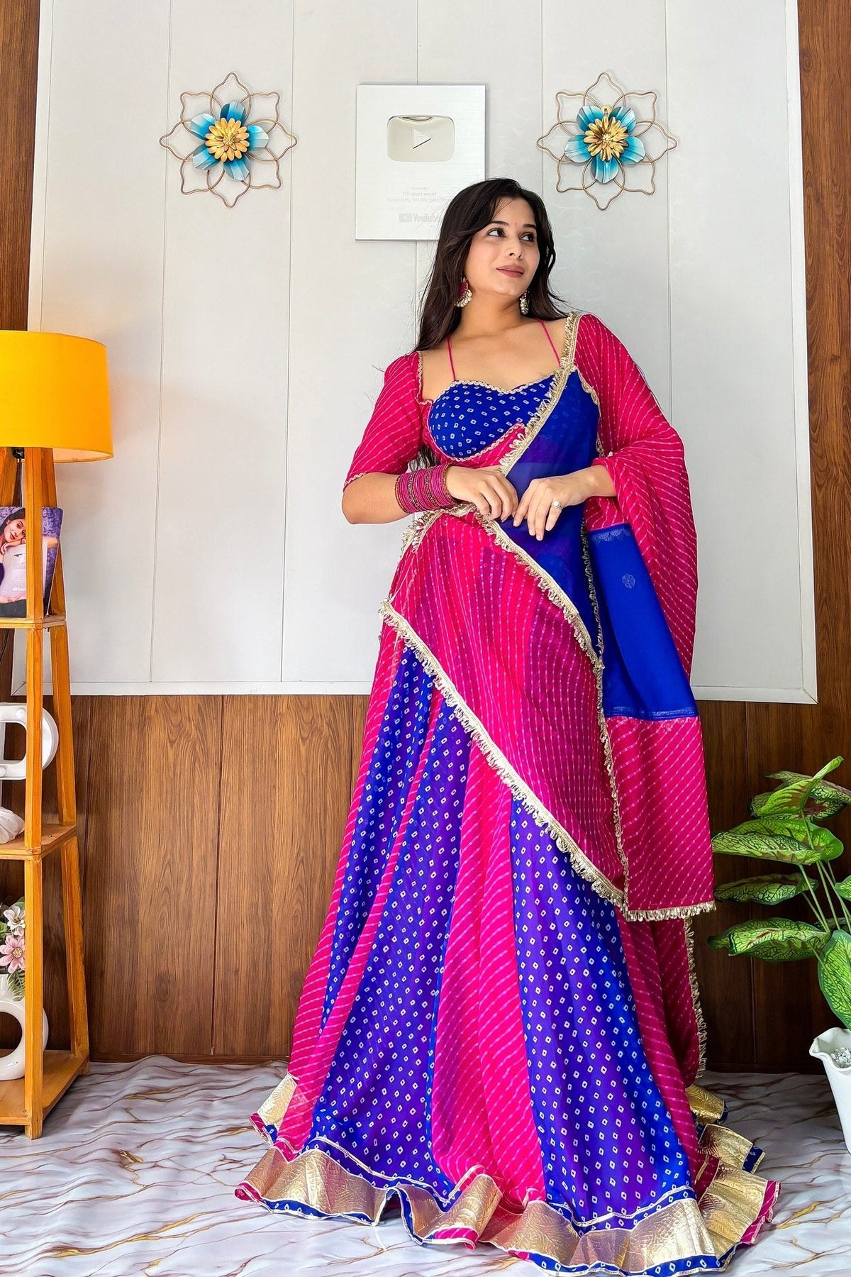 A woman models a traditional Indian lehenga choli dress in vibrant pink and blue with gold borders, standing in a decorated room with a wooden floor, plants, and wall decor.
