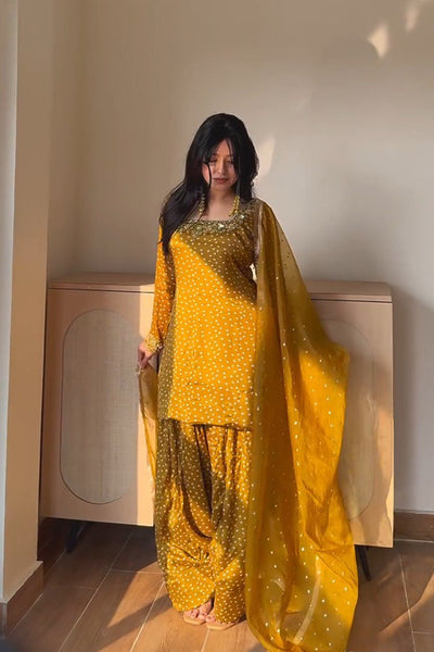 A woman wearing a bright yellow traditional outfit with a matching sheer dupatta, standing in a sunlit indoor room with a wooden cabinet behind her.