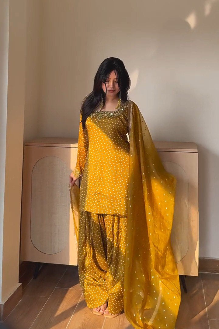 A woman wearing a bright yellow traditional outfit with a matching sheer dupatta, standing in a sunlit indoor room with a wooden cabinet behind her.