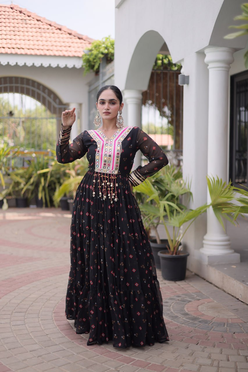 A woman stands outdoors in a courtyard wearing a long black embroidered dress with pink and white detailing on the bodice and scattered floral motifs throughout the skirt. The setting includes white columns, arches, potted plants, and a tiled roof in the background.