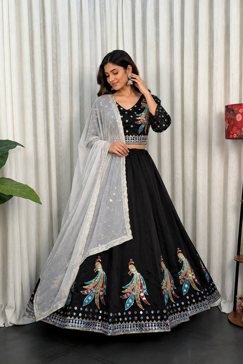 A woman models a black Indian ethnic outfit with a flared skirt, embroidered peacock motifs, a matching embellished blouse, and a sheer white dupatta, posed in front of a white curtain backdrop.