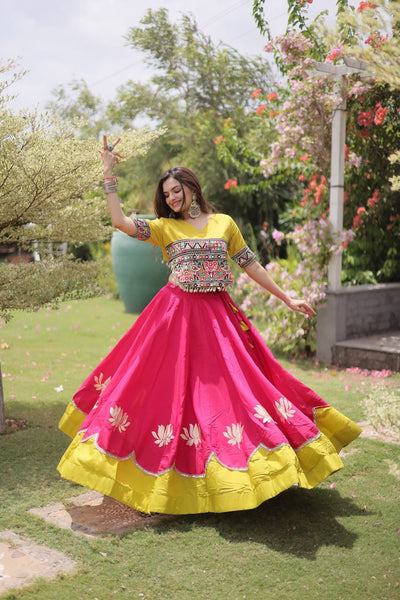 A woman outdoors in a lush garden wearing a bright pink and yellow traditional outfit with floral embroidery and a flared skirt, captured mid-twirl.