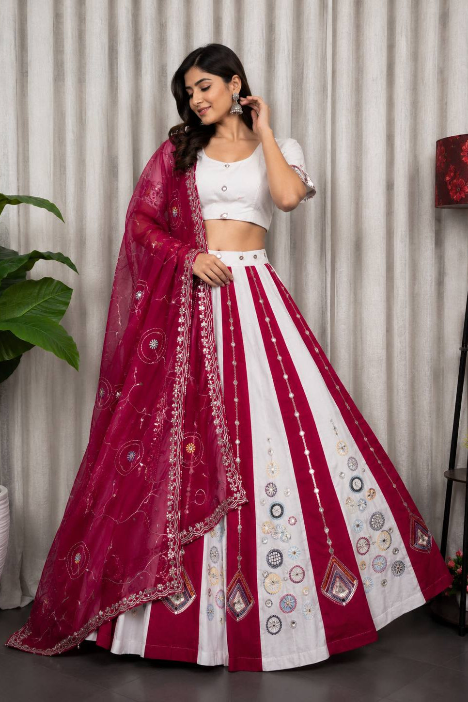 A woman models a traditional Indian lehenga choli set. The outfit features a white skirt with vertical magenta stripes and circular embroidered motifs, paired with a matching white crop top and a sheer magenta dupatta decorated with intricate gold and white embroidery.
