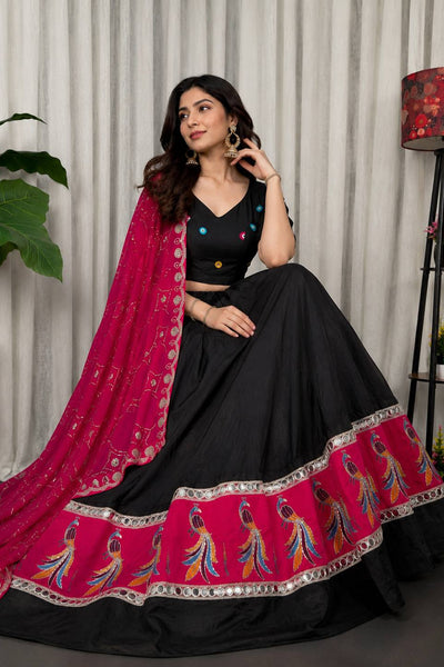 A woman models a black and magenta lehenga choli set with detailed embroidery and mirror work, posed in front of a neutral curtain backdrop.