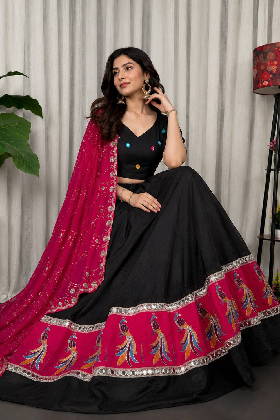 A woman models a black and magenta lehenga choli set with detailed embroidery and mirror work, posed in front of a neutral curtain backdrop.