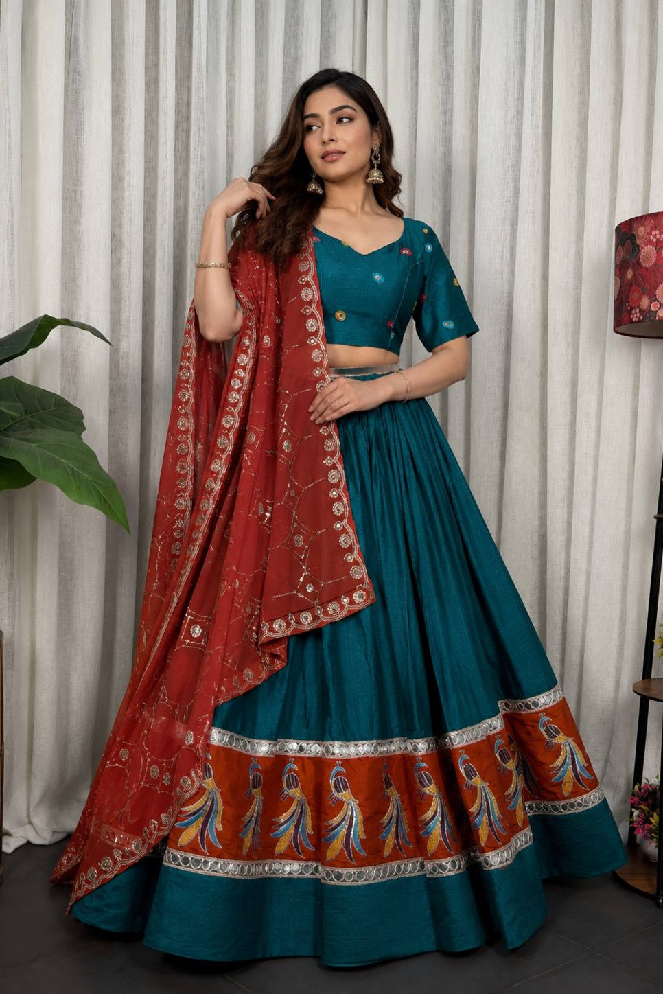 A woman models a traditional Indian lehenga choli set. The outfit features a deep teal blue skirt (lehenga) and blouse (choli) with gold button details, paired with a red dupatta (scarf) adorned with intricate gold and silver zari embroidery and a matching orange border print.