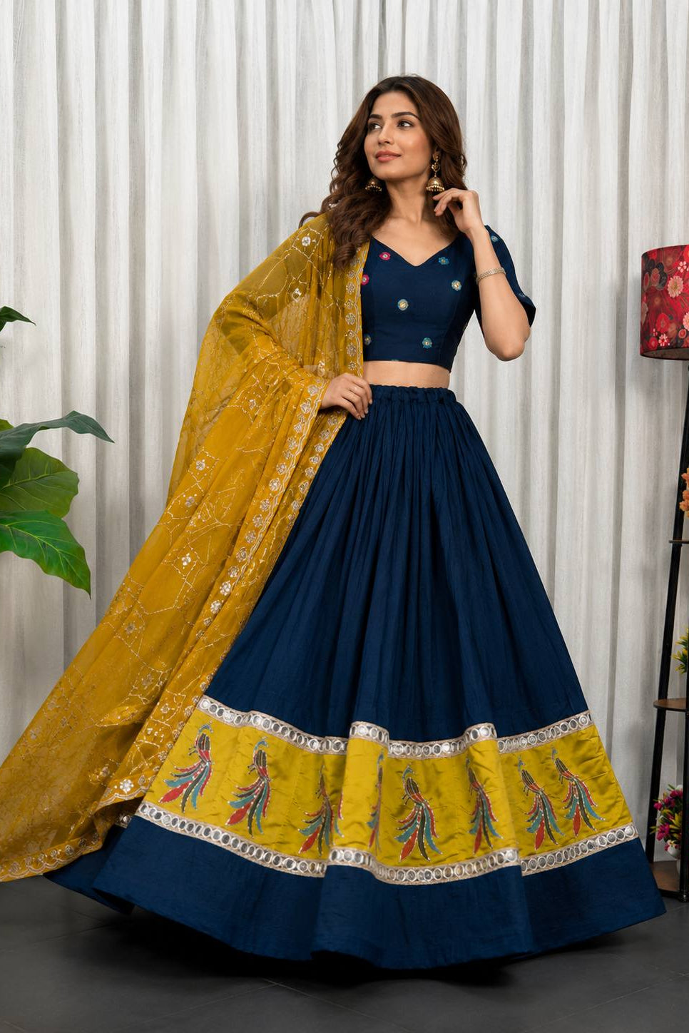 A woman models a navy blue and mustard yellow traditional Indian outfit with a matching dupatta. The outfit features a navy blue skirt and blouse with gold embroidery, and a wide yellow border on the skirt with colorful printed motifs.