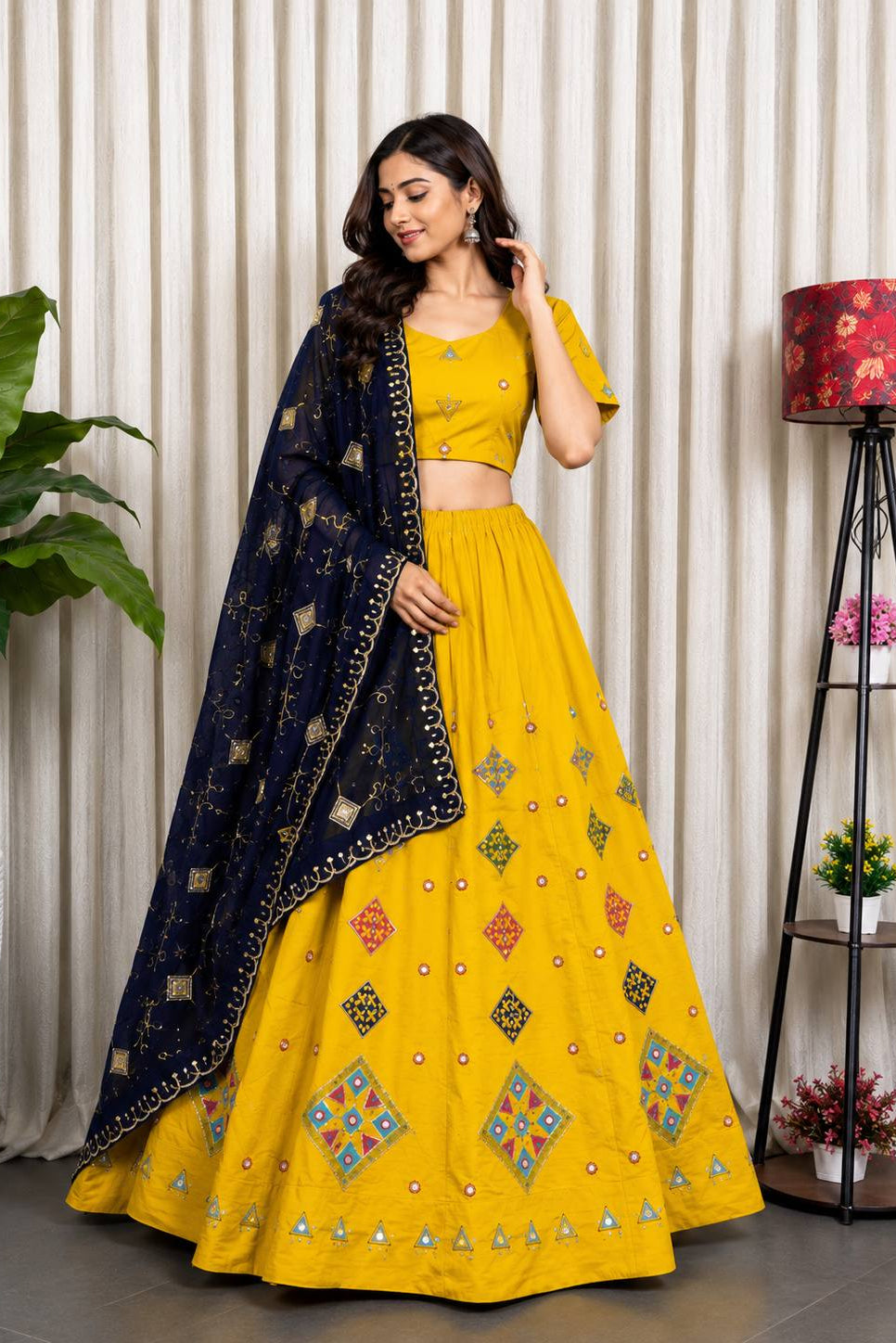 A woman models a traditional Indian lehenga choli set in a studio setting. The outfit consists of a bright yellow skirt (lehenga) with colorful geometric embroidery, a matching yellow crop top (choli), and a navy blue dupatta with gold and multicolor embroidery.