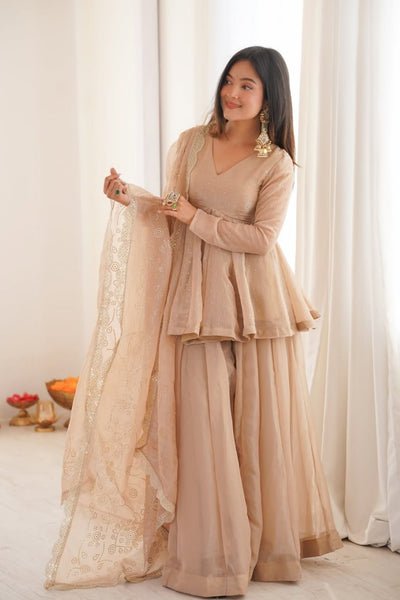 Woman in a beige traditional outfit holding a dupatta against a white curtain background
