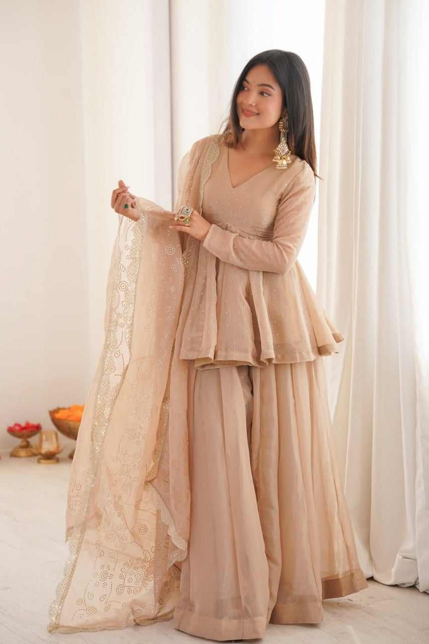 Woman in a beige traditional outfit holding a dupatta against a white curtain background