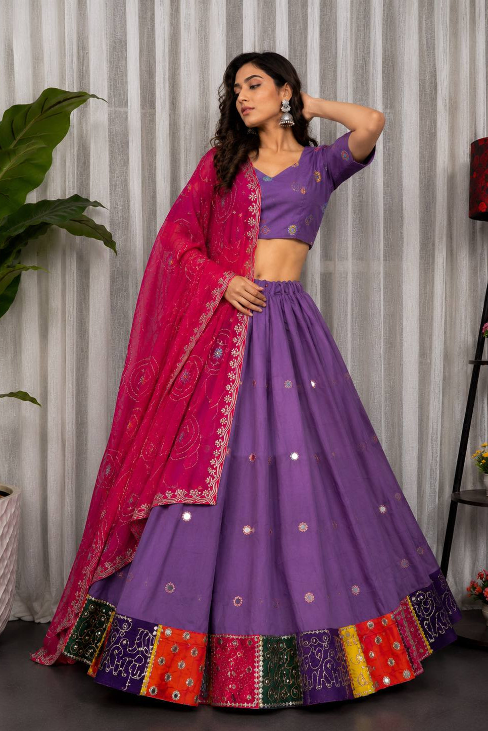 A woman models a traditional Indian lehenga choli set. The outfit features a purple flared skirt (lehenga) with small silver mirror work details, a matching purple short-sleeve blouse, and a bright pink dupatta with intricate silver embroidery and mirror work.