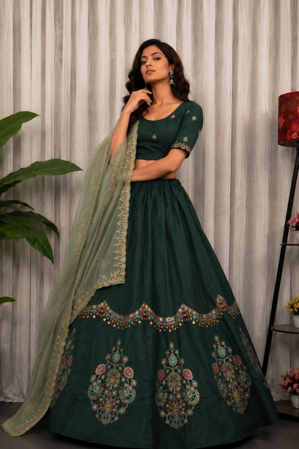 A woman models a dark green Indian ethnic wear set featuring a flared lehenga skirt with colorful floral embroidery, a matching embroidered blouse, and a sheer dupatta, set against a neutral curtain backdrop.