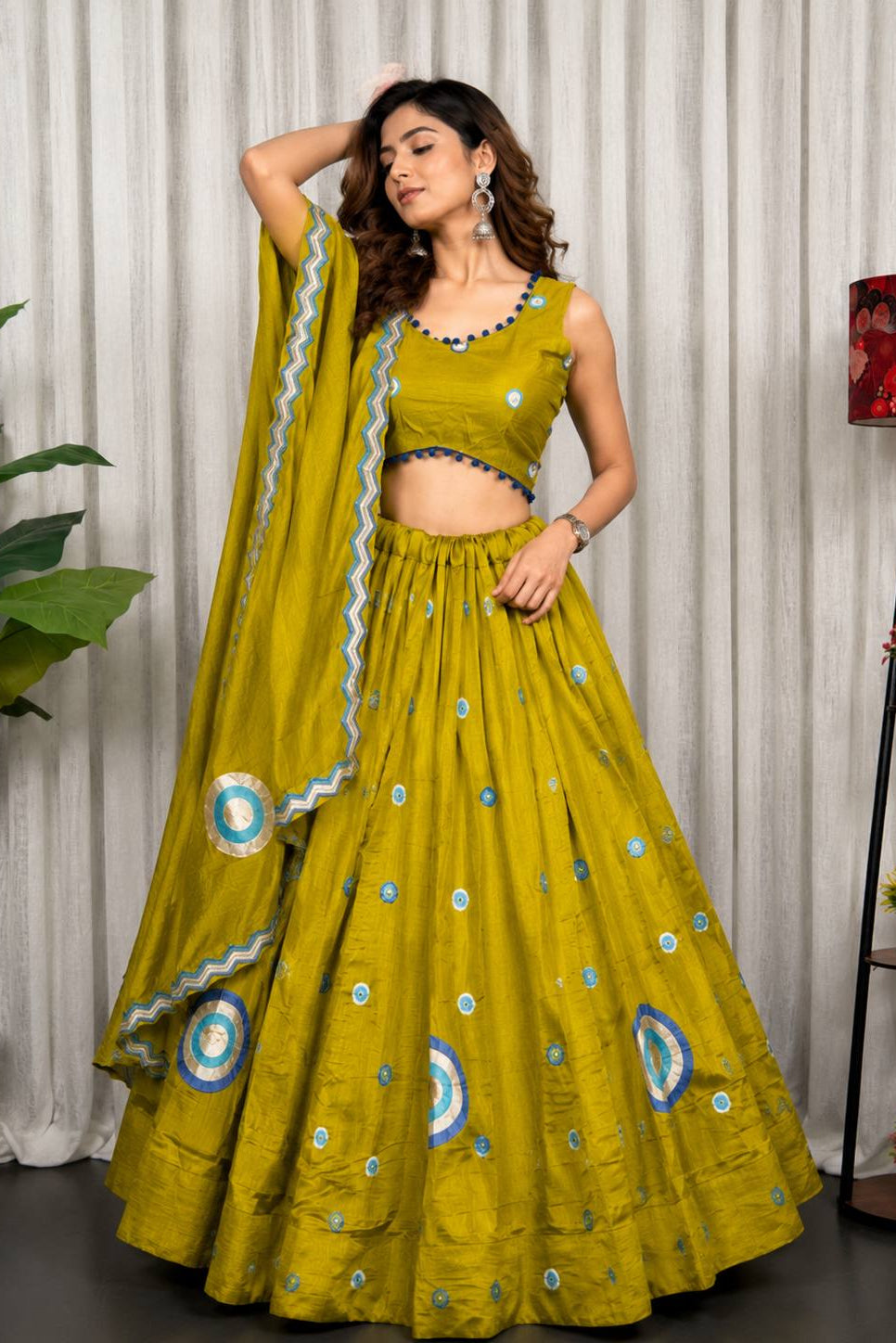 A woman models a traditional Indian lehenga choli set in olive green with blue and white embroidered circular motifs and a matching dupatta, set against a neutral indoor backdrop with curtains and decorative plants.