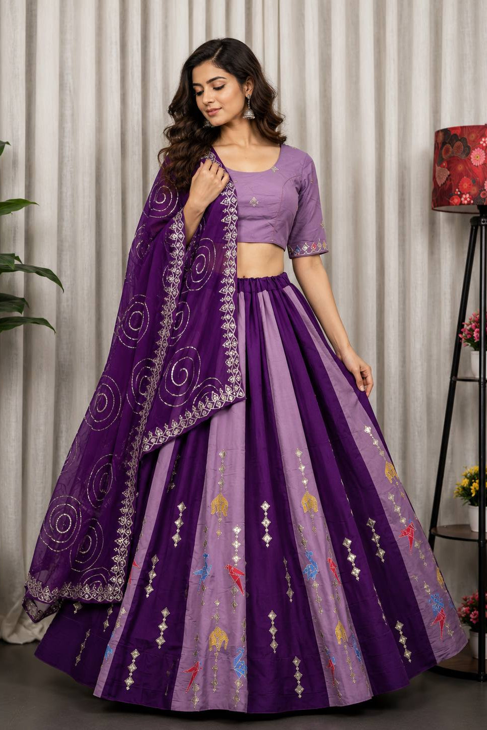 A woman models a traditional Indian lehenga choli set in shades of purple, featuring a lavender purple blouse, a matching purple flared skirt with colorful embroidery, and a sheer purple dupatta with silver mirror work.