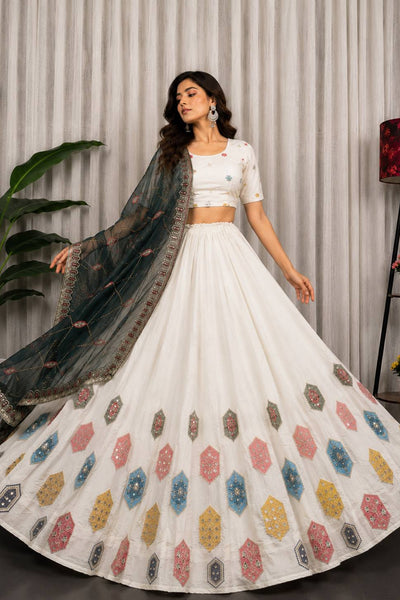 A woman models a white traditional lehenga choli set with a dark teal dupatta. The skirt features colorful geometric embroidered motifs in pink, blue, yellow, and brown.