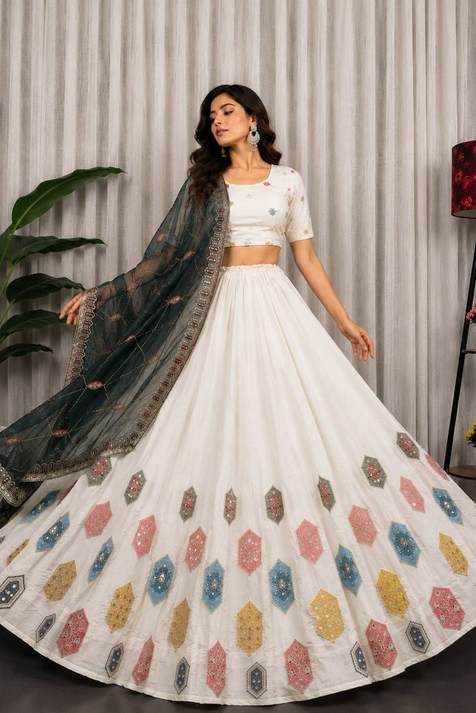 A woman models a white traditional lehenga choli set with a dark teal dupatta. The skirt features colorful geometric embroidered motifs in pink, blue, yellow, and brown.