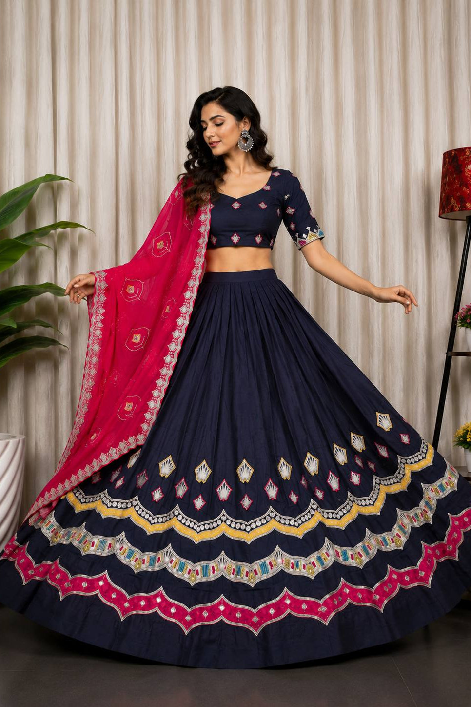 A woman models a navy blue Indian ethnic lehenga choli set with a bright pink dupatta. The skirt features intricate multicolor embroidery along the hem, and the blouse has embroidered sleeves.