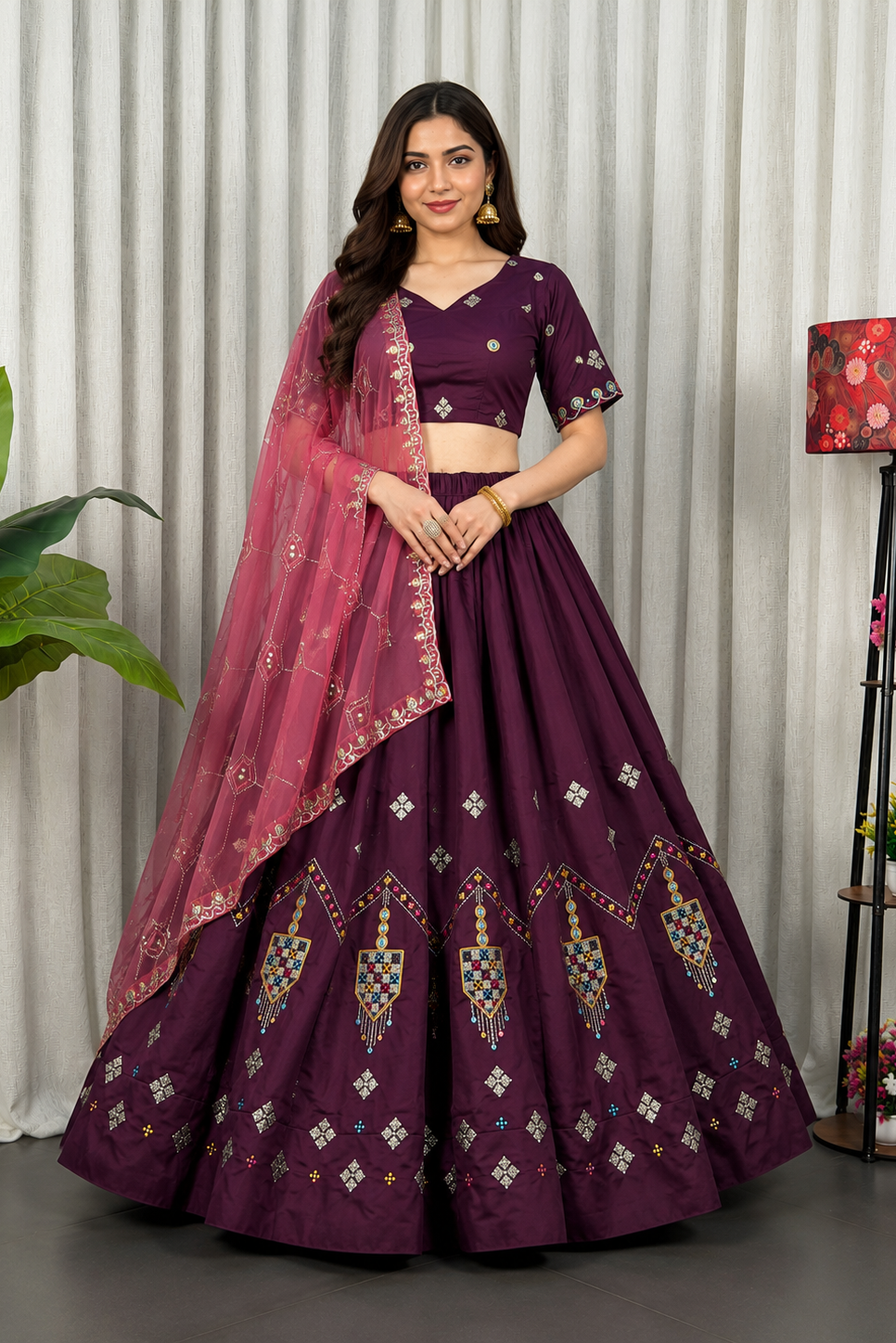 A woman models a traditional Indian lehenga choli set in deep purple, featuring a flared skirt with geometric embroidery, a matching embroidered blouse, and a sheer dupatta in a contrasting pinkish-purple shade.