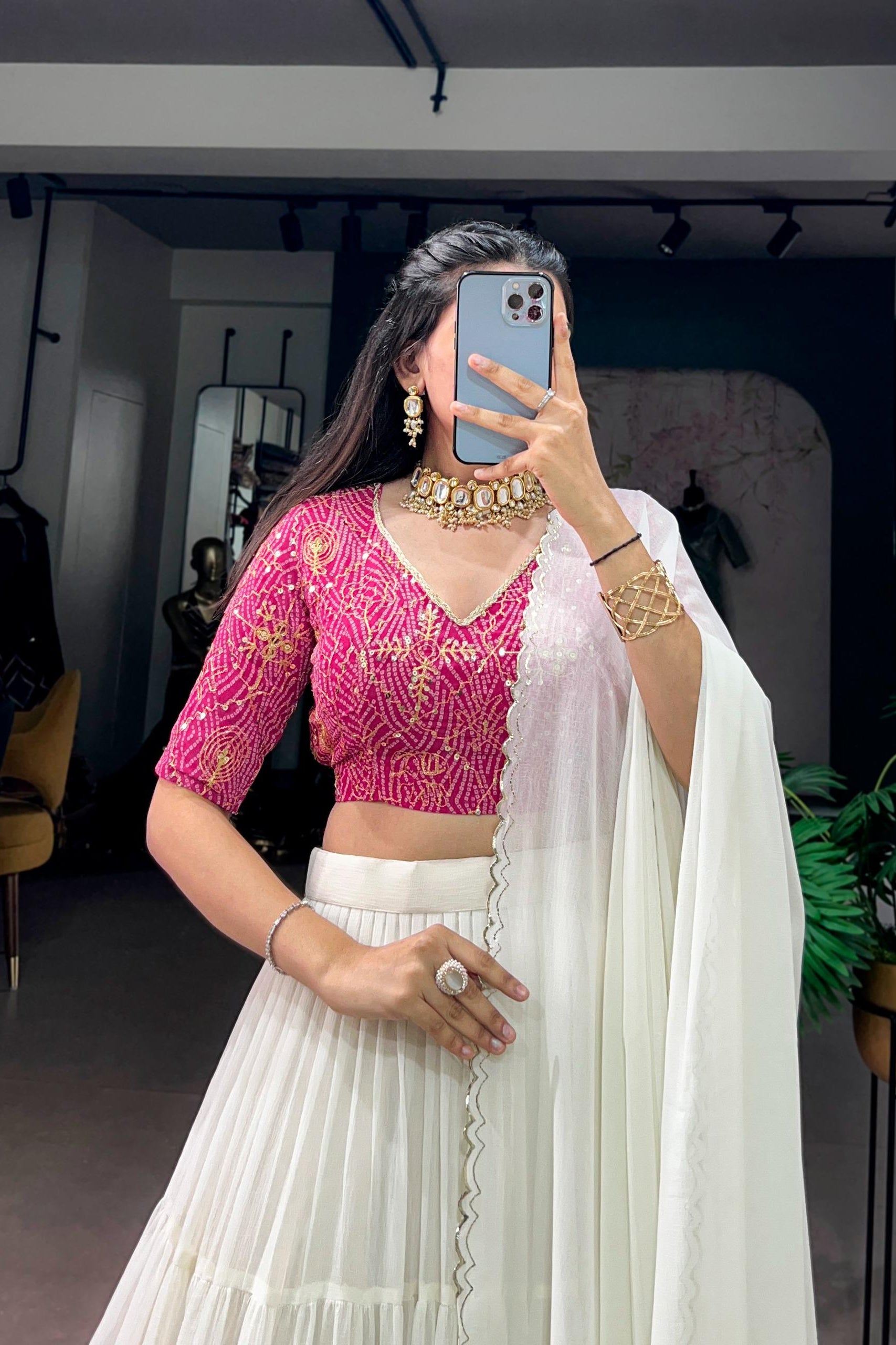 A woman takes a mirror selfie wearing a traditional Indian outfit consisting of a bright pink embroidered crop top, a white pleated skirt, and a white dupatta draped over her shoulder. She is accessorized with gold jewelry, including a statement necklace, bracelet, and rings.