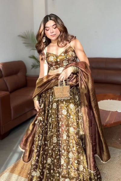 A woman models a brown and gold floral lehenga choli set with a matching dupatta in a living room setting.