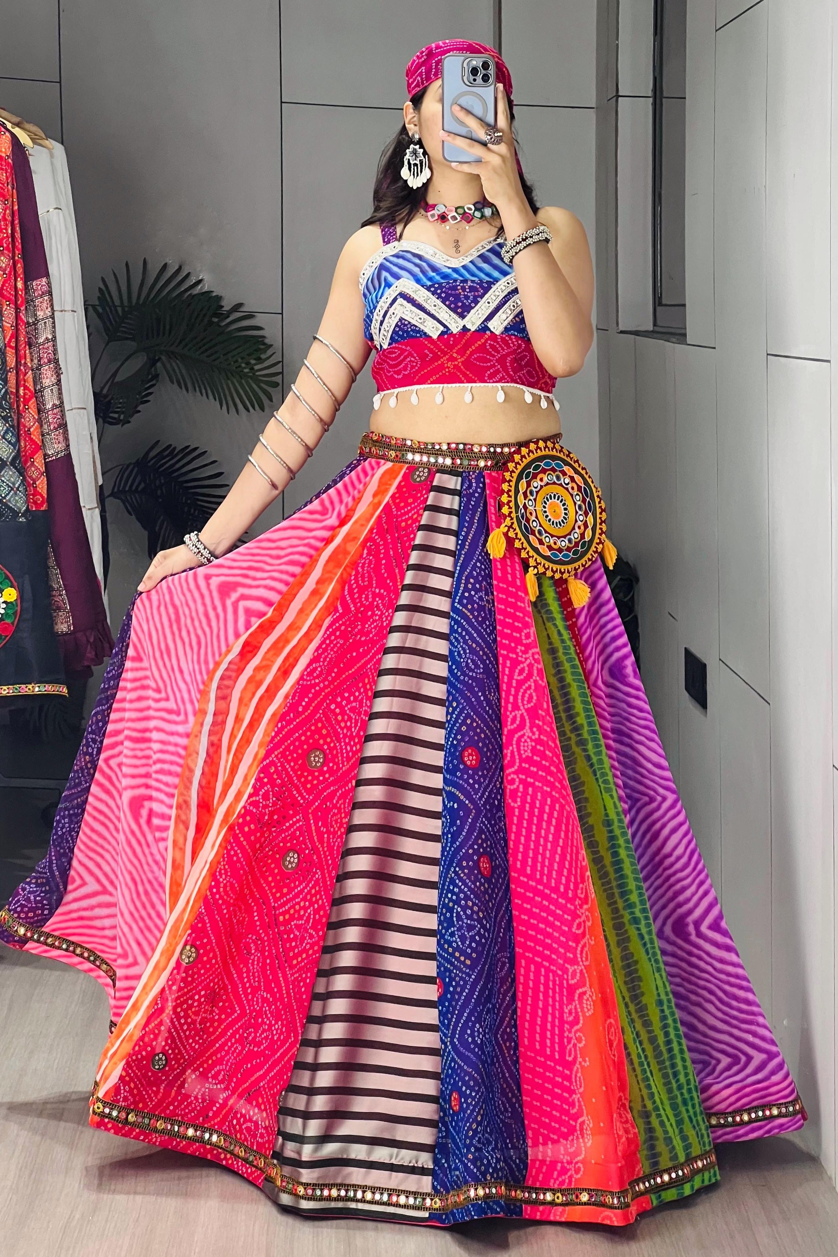A woman takes a mirror selfie wearing a vibrant, multicolored traditional Indian lehenga choli outfit. The skirt features multiple panels of different patterns and colors including pink, purple, blue, orange, and green, with a matching headpiece and jewelry.