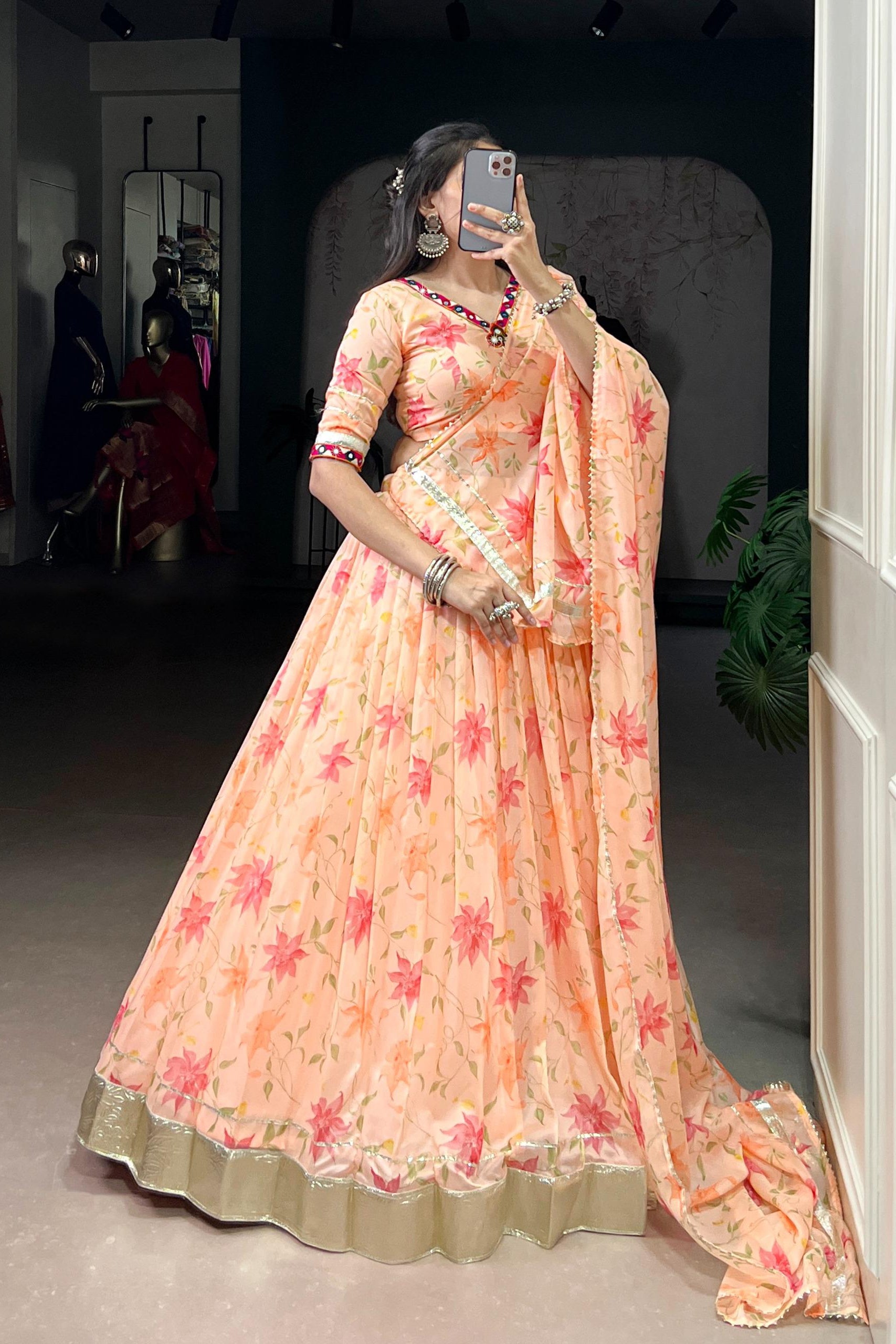 Woman in a peach floral saree taking a mirror selfie indoors.