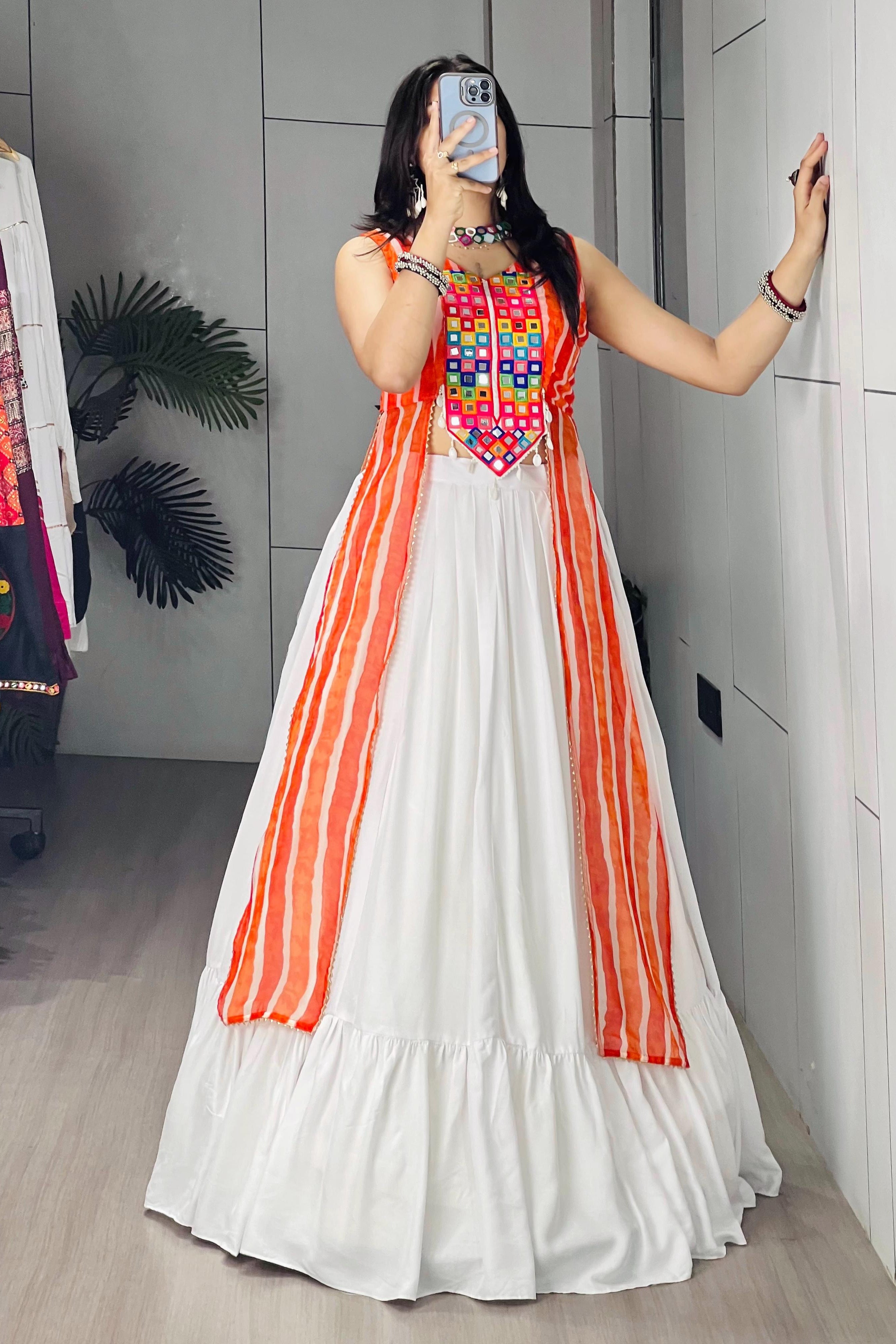 A woman takes a mirror selfie in a boutique wearing a white A-line dress with vibrant orange vertical stripes and a colorful embroidered bodice. She is standing in a fitting room with a gray paneled wall and a clothing rack visible on the left.