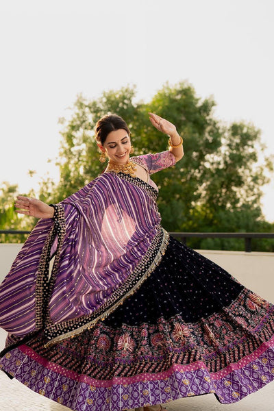 Woman in a traditional outfit with a purple and black patterned dress, standing outdoors with greenery in the background.