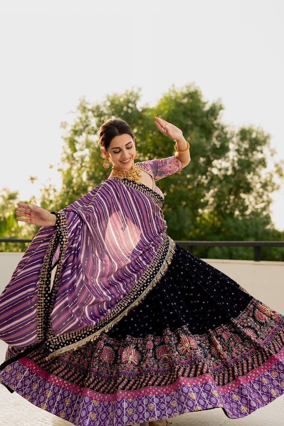 Woman in a traditional outfit with a purple and black patterned dress, standing outdoors with greenery in the background.